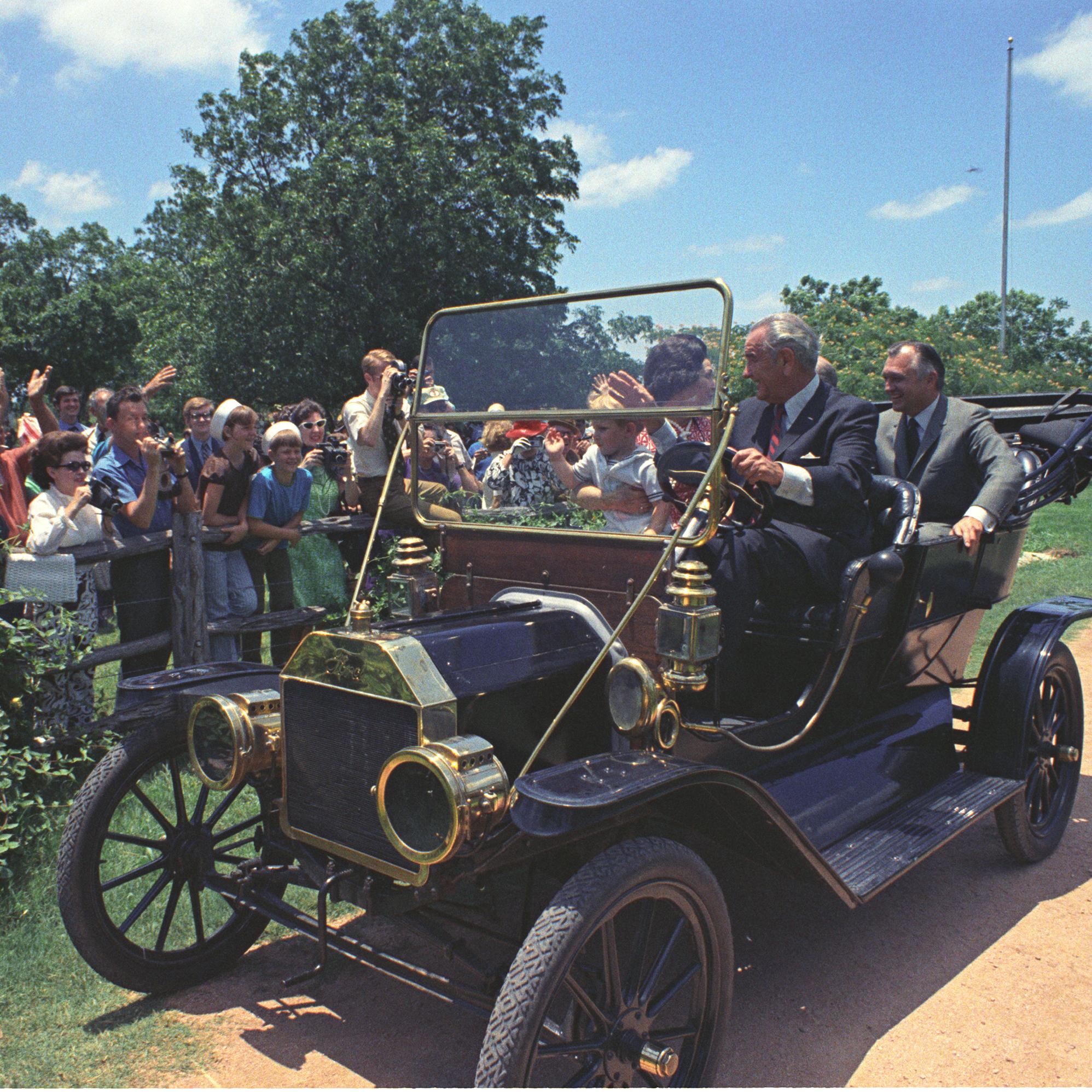 President Lyndon B. Johnson driving his Model-T Ford down a dirt road, waving to crowd. Lady Bird Johnson, grandson Lyn Nugent and others ride along.